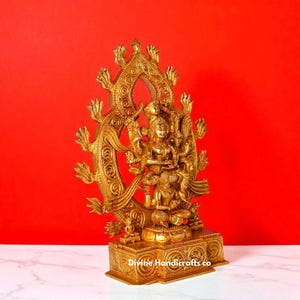 Puede incluir: Una estatua dorada de una deidad con m&uacute;ltiples brazos y figuras, sobre un fondo rojo. La estatua presenta detalles intrincados y est&aacute; montada sobre una base decorativa. El texto "Divine Handicrafts co" es visible en la base.
