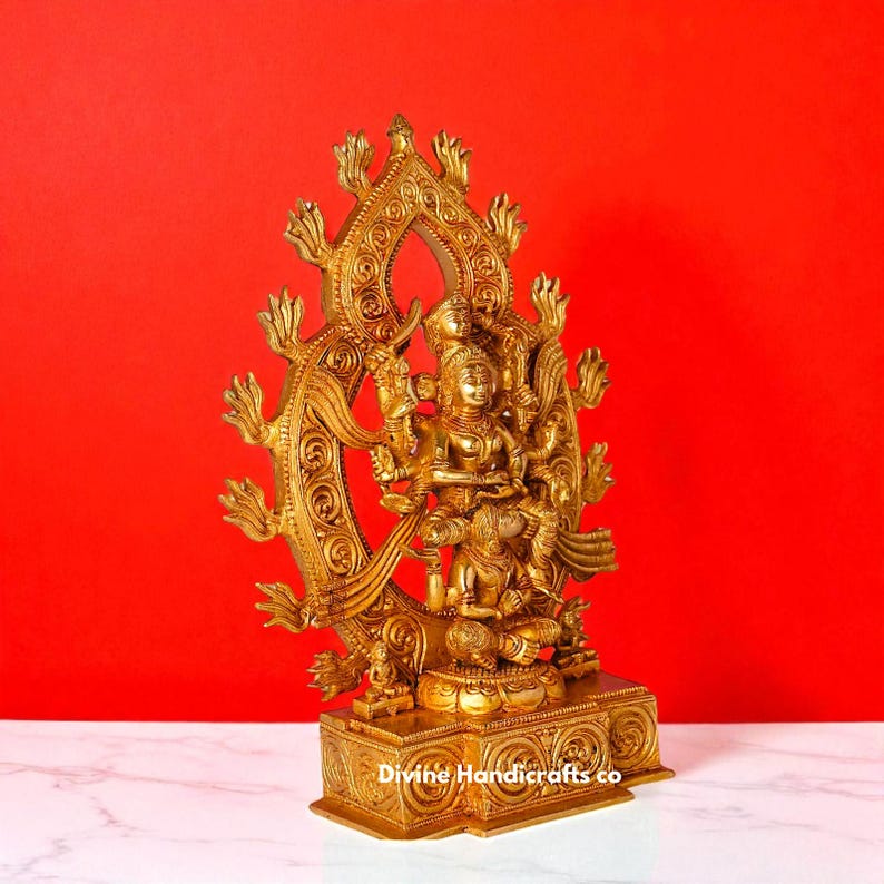 Puede incluir: Una estatua dorada de una deidad con m&uacute;ltiples brazos y figuras, sobre un fondo rojo. La estatua presenta detalles intrincados y est&aacute; montada sobre una base decorativa. El texto "Divine Handicrafts co" es visible en la base.