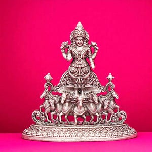 May include: A silver-colored statue of a deity, adorned with intricate details, stands on a decorative base. The statue features a central figure with multiple arms, surrounded by horses. The background is a vibrant pink.