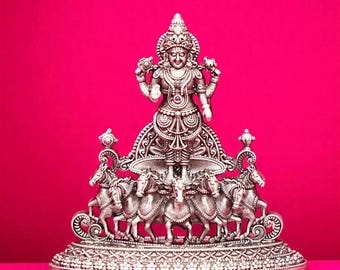 May include: A silver-colored statue of a deity, adorned with intricate details, stands on a decorative base. The statue features a central figure with multiple arms, surrounded by horses. The background is a vibrant pink.