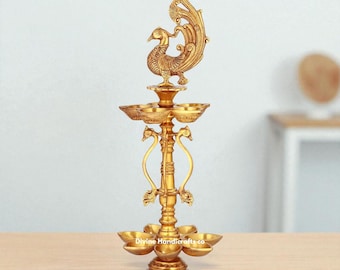 May include: A golden brass oil lamp with a peacock design. The lamp has multiple tiers of bowls for oil and a decorative peacock at the top. The base is a flower shape. The text "Divine Handicrafts.co" is visible.