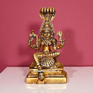 Puede incluir: Estatua dorada de una deidad sentada, con múltiples brazos y una capucha de cobra. La estatua está intrincadamente detallada, con ropa y joyas ornamentadas. La base de la estatua tiene una cara tallada. El fondo es rosa liso.