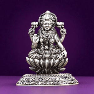 May include: Silver-colored statue of a seated deity, adorned with intricate jewelry and a halo. The figure is seated on a lotus flower base, with a decorative rectangular pedestal. The background is a solid purple.