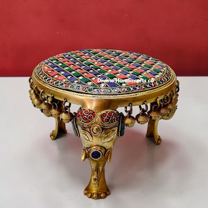 May include: A brass decorative table with a mosaic top featuring red, green, blue, and white stones. The table has elephant-shaped legs and small bells hanging from the rim. The table is a unique piece of home decor.