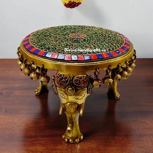 May include: A brass decorative table with an elephant-head base. The round top is inlaid with green, red, and blue stones. Small bells hang from the table's edge. The text "Divine Handicraft co" is visible on the top.