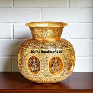 May include: A gold-colored, ornate vase with intricate floral and deity carvings. The vase has a wide, flared rim and a rounded body. The text "Divine Handicrafts co" is visible on the vase.