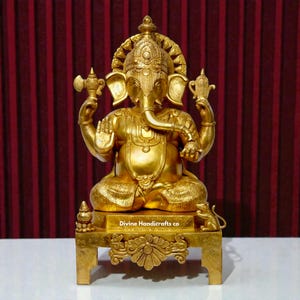 May include: A gold-colored statue of the Hindu deity Ganesha, seated in a meditative pose. The statue features intricate details, including a large elephant head, multiple arms, and ornate jewelry. The statue is on a decorative base with the text "Divine Handicrafts co."