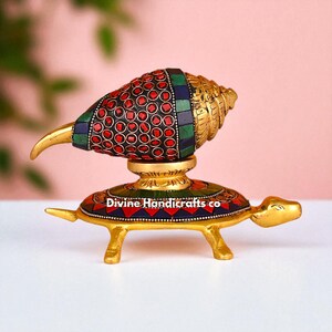 May include: A gold-colored decorative conch shell with red, green, and blue accents, resting on a turtle-shaped base. The base is also gold and features colorful mosaic details. The text "Divine Handicrafts co" is visible on the base.