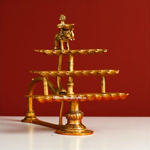May include: A three-tiered brass oil lamp with a figure riding an animal on top. The lamp has multiple holders for oil and wicks. The base is ornate, and the overall design is intricate and detailed.