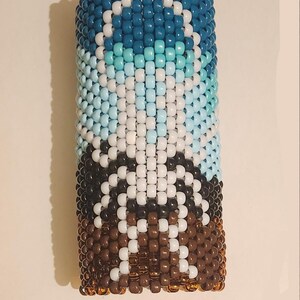 May include: A cylindrical cuff bracelet crafted from small plastic pony beads arranged in a vertical pattern. The design features a gradient of dark blue, light blue, and white beads at the top, transitioning into brown and white beads at the base.
