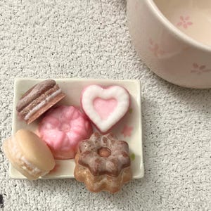 May include: A small rectangular plate holds six decorative soaps in the shape of a macaron, a layered pastry, a heart, a flower, and a pink, round shape. A pink teacup with floral designs is in the background.