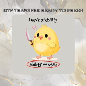May include: A DTF transfer featuring a yellow cartoon chick holding a knife. The text "i have stability" is above the chick, and "ability to stab" is written below. The background is light gray with gold accents.