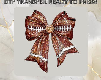 May include: A DTF transfer design featuring a glittery brown bow with white football stitching and accents. The bow is set against a light gray background with the text "DTF TRANSFER READY TO PRESS" at the top.