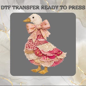 Peut inclure: Un transfert DTF READY TO PRESS représentant un canard orné de pièces de tissu superposées dans des tons de rose, crème et rouge. Le canard porte un grand nœud rose avec des accents dorés, ainsi qu'un bec et des pattes à paillettes dorées.