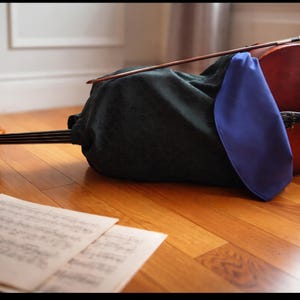 Peut inclure: Un violoncelle avec un corps en bois foncé et un archet repose sur un sol en bois. Une housse vert foncé et un tissu bleu recouvrent partiellement l'instrument. Des partitions sont au premier plan.