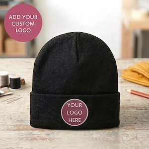 Puede incluir: Un gorro de punto negro liso con un parche circular en el dobladillo frontal. El parche es de color malva con el texto blanco "YOUR LOGO HERE" centrado. El gorro presenta un diseño clásico con dobladillo y una textura de punto visible.