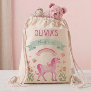 Może przedstawiać: Beżowa torba do zabawy ze sznurkiem z różowym napisem "Olivia's". Torba ma różowy wzór jednorożca, tęczy i kwiatów. Mała lalka i różowy miś są widoczne na górze.