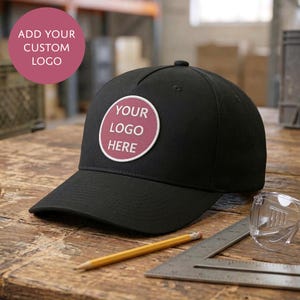 以下が含まれることがあります： 前面パネルにマルーン色の背景で「YOUR LOGO HERE」という白い文字が記された円形のパッチが付いた黒い野球帽。帽子は木製の表面に置かれた黄色い鉛筆と金属製の差し金の近くに配置されています。