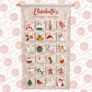 Peut inclure: Un calendrier de l'Avent en tissu crème suspendu à une tige en bois. Il porte l'inscription Elizabeth's Christmas Countdown et vingt-quatre poches numérotées, chacune ornée d'illustrations de Noël colorées comme un bonhomme de neige, le Père Noël et des cloches.