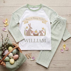 Pode incluir: Conjunto de pijama de Páscoa para crianças. A parte superior branca tem mangas listradas verdes e um gráfico com o texto "Have a Hoppy Easter", coelhos e uma cesta de ovos. O nome "WILLIAM" é impresso abaixo. As calças são listradas em verde.