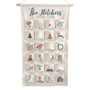 May include: A cream-coloured Christmas advent calendar with 24 numbered pockets, each featuring a festive illustration. The calendar is labelled "The Fletchers Christmas Calendar" in green script and hangs from a wooden dowel.