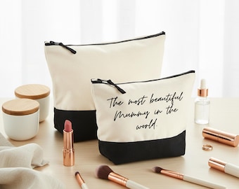 Puede incluir: Dos neceseres de lona crema con bases negras y cremalleras. La bolsa más pequeña tiene el texto "The most beautiful Mummy in the world". Se muestran brochas de maquillaje, pintalabios y otros productos de belleza.