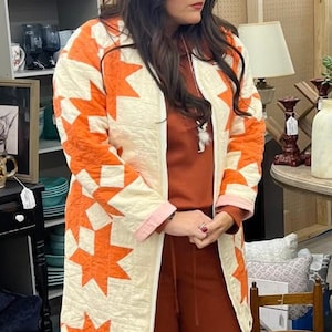May include: A long, quilted coat with a star pattern in orange and white. The coat is worn over a rust-colored outfit. The coat has a cream-colored background with orange star shapes. The coat has a pink trim.