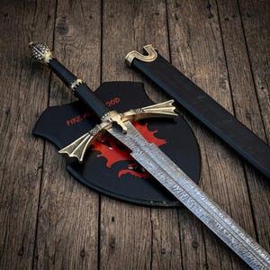 May include: A Damascus steel sword with a black handle and ornate brass crossguard and pommel. The sword rests on a black shield plaque featuring a red dragon graphic and the text FIRE AND BLOOD, accompanied by a matching black scabbard.