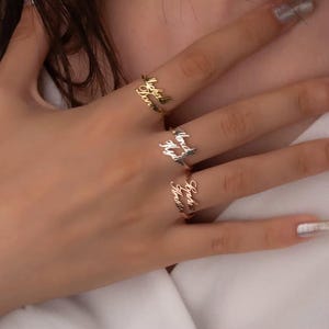 Può includere: Tre anelli personalizzati con nomi in oro, argento e oro rosa. Ogni anello presenta un nome diverso in un delicato carattere di scrittura. Gli anelli sono esposti su una mano, mettendo in risalto il design e lo stile dei gioielli.