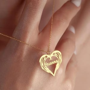 Puede incluir: Un collar dorado con un colgante en forma de coraz&oacute;n. El colgante presenta el nombre "Brianna" en escritura cursiva, rodeado por un dise&ntilde;o de alas. El collar est&aacute; en una cadena delicada.