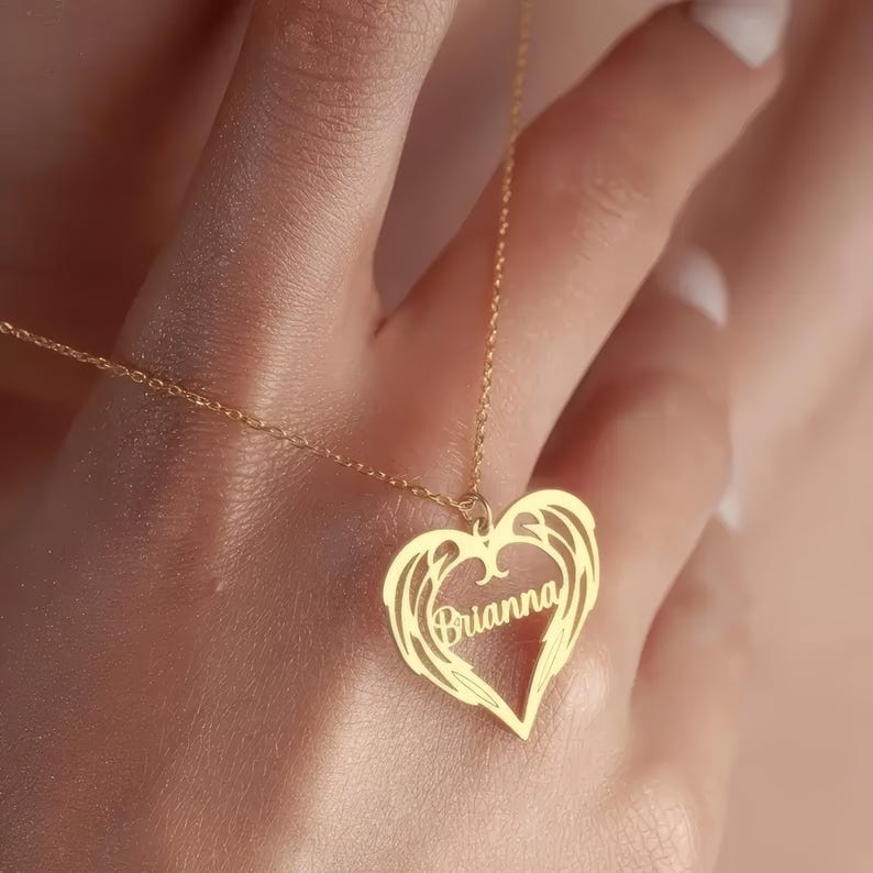 Puede incluir: Un collar dorado con un colgante en forma de coraz&oacute;n. El colgante presenta el nombre "Brianna" en escritura cursiva, rodeado por un dise&ntilde;o de alas. El collar est&aacute; en una cadena delicada.