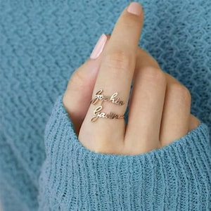 May include: A gold-coloured ring with two bands, each bearing a name in cursive script. The top band reads "Sophia," and the bottom band reads "Gavin." The ring is worn on a finger, set against a blue background.