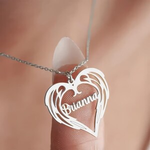 Puede incluir: Un collar plateado con un colgante en forma de coraz&oacute;n con dise&ntilde;os de alas y el nombre "Brianna". El colgante cuelga de una delicada cadena. El fondo est&aacute; borroso, enfocando la atenci&oacute;n en el collar.