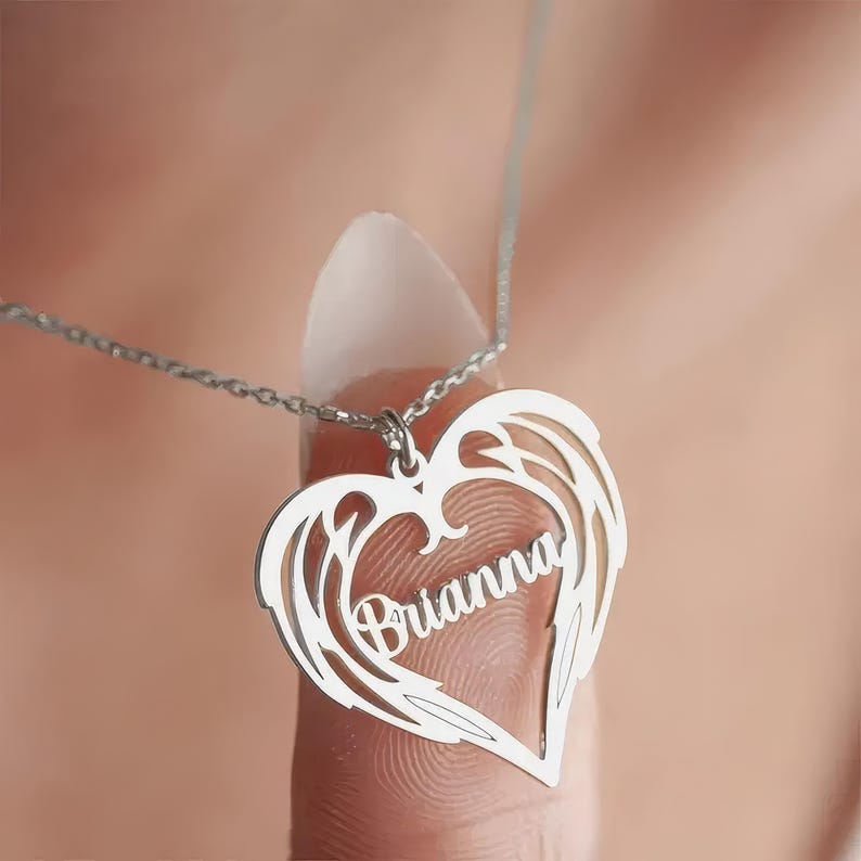 Puede incluir: Un collar plateado con un colgante en forma de coraz&oacute;n con dise&ntilde;os de alas y el nombre "Brianna". El colgante cuelga de una delicada cadena. El fondo est&aacute; borroso, enfocando la atenci&oacute;n en el collar.