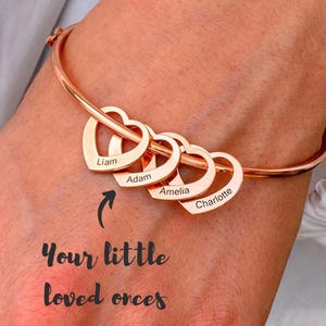 Puede incluir: Pulsera de brazalete de oro rosa con cuatro colgantes en forma de corazón. Cada corazón está grabado con un nombre: Liam, Adam, Amelia y Charlotte. La pulsera se muestra en una muñeca.
