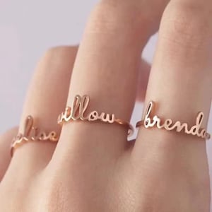 May include: Three rose gold rings, each spelling a name in cursive: "Lisa", "Willow", and "Brenda". The rings are shown on a person's fingers, highlighting the personalised design. The rings are a delicate piece of jewellery.