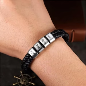 Op de afbeelding: Zwart gevlochten leren armband met zilveren naamkralen. De armband heeft de namen "Logan", "Avery", "Mason" en "Luna" gegraveerd op de kralen. De armband wordt om een pols gedragen.