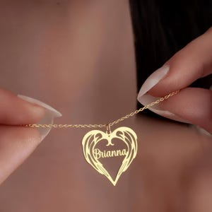 Puede incluir: Un collar dorado con un colgante en forma de coraz&oacute;n. El colgante presenta un dise&ntilde;o de alas y el nombre "Brianna" en escritura cursiva. El collar se sostiene, mostrando la fina cadena y los detalles del colgante.