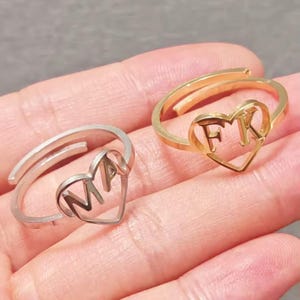 May include: Two adjustable rings. One is silver with "MA" in a heart-shaped cutout, the other gold with "FK". The rings are designed to be worn on a finger. The rings are made of metal.