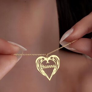 Puede incluir: Un collar dorado con un colgante en forma de coraz&oacute;n. El colgante presenta un dise&ntilde;o de alas y el nombre "Brianna". El collar se sostiene, mostrando la fina cadena y el detalle del colgante.