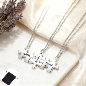 Könnte beinhalten: Drei silberne Puzzleteil-Halsketten, jeweils mit dem Wort "NAME" graviert. Die Halsketten werden auf einer weißen Oberfläche präsentiert, mit einem kleinen schwarzen Beutel unten links und Lavendelzweigen oben links.