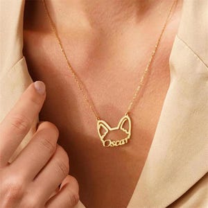 Pode incluir: Um colar dourado com um pingente de silhueta de cachorro com o nome "Oscar". O pingente é suspenso por uma corrente fina. O colar é exibido em um fundo bege.
