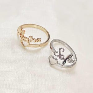May include: Two personalised rings. One is gold-toned with the name "Karina" and a heart-shaped accent. The other is silver-toned with the name "Sofia" and a heart-shaped accent. Both rings are on a white surface.