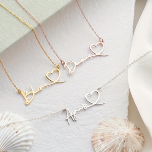 May include: Four personalised name necklaces in gold, rose gold, and silver. Each necklace features a name in a delicate script font, with a heart design. The necklaces are displayed on a white, textured surface.