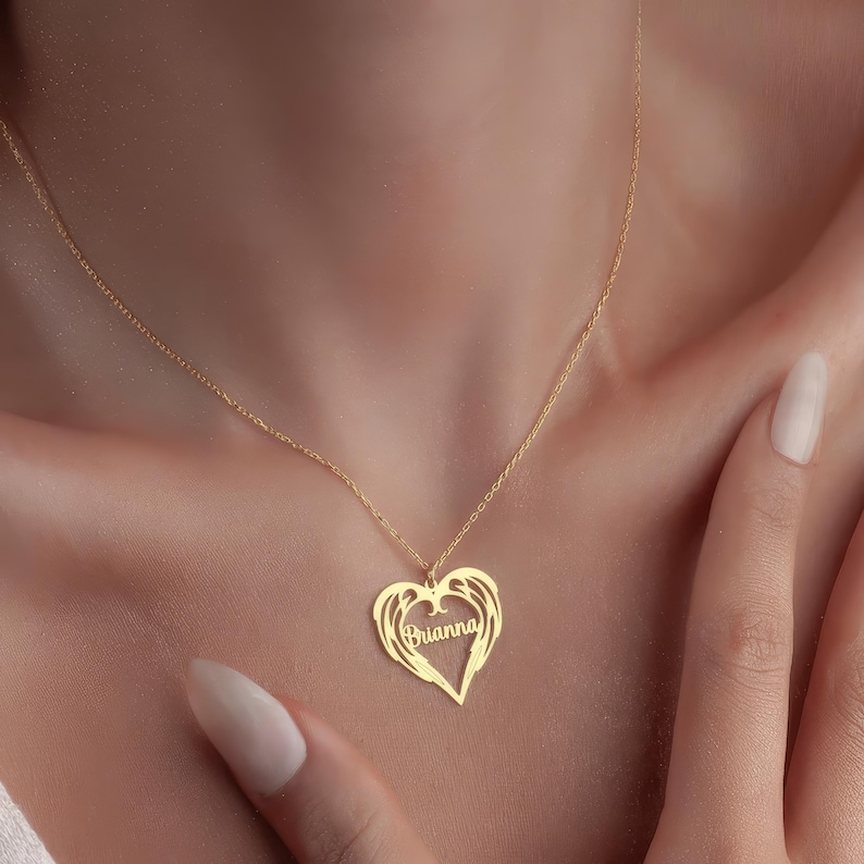 Puede incluir: Un collar dorado con un colgante en forma de coraz&oacute;n. El colgante tiene el nombre "Brianna" en escritura cursiva dentro de un coraz&oacute;n con detalles de alas. El collar se lleva alrededor del cuello.