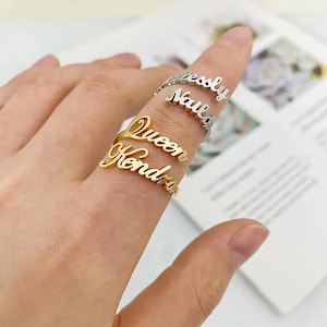 以下が含まれることがあります： 金と銀の名前入りリングのコレクション。リングには、金色の「Queen」と「Kendra」、銀色の「Jessly」と「Naula」の名前が刻まれています。指にリングがはめられています。