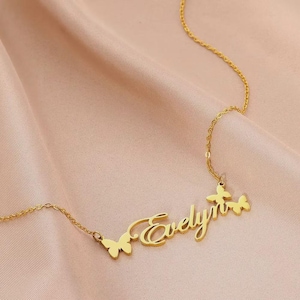 Puede incluir: Un collar dorado con el nombre "Evelyn" en escritura cursiva. El collar presenta detalles de mariposas a cada lado del nombre y al final de la cadena. El collar se muestra sobre una tela rosa.