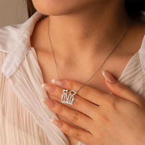 May include: A silver necklace featuring a cat and a dog pendant. The pendant has the names "Samantha" and "Benjamin" engraved on it. The necklace is worn by a person wearing a white blouse.