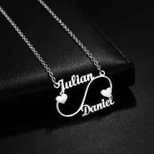 Puede incluir: Un collar plateado con los nombres "Julian" y "Daniel" en escritura cursiva, conectados por un símbolo de infinito. Pequeños dijes en forma de corazón están colocados cerca de cada nombre. El collar se muestra sobre una superficie negra.