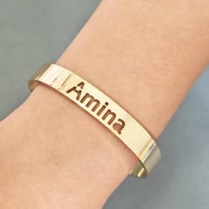 Puede incluir: Una pulsera de brazalete dorada con el nombre "Amina" recortado en el centro. La pulsera es lisa y brillante, y se lleva en la muñeca. El fondo es de color gris liso.